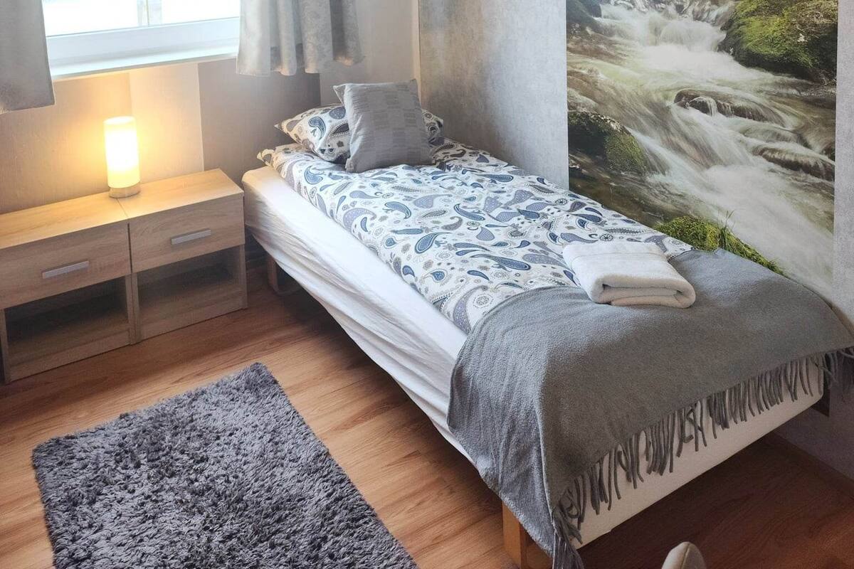 A bedroom with a bed, a nightstand, a lamp, a window with curtains, and a large wall mural of a waterfall.