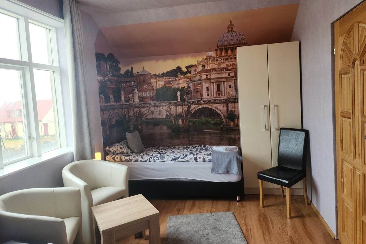 A bedroom with a bed, a chair, a desk, a wardrobe, and a large mural of a cityscape.