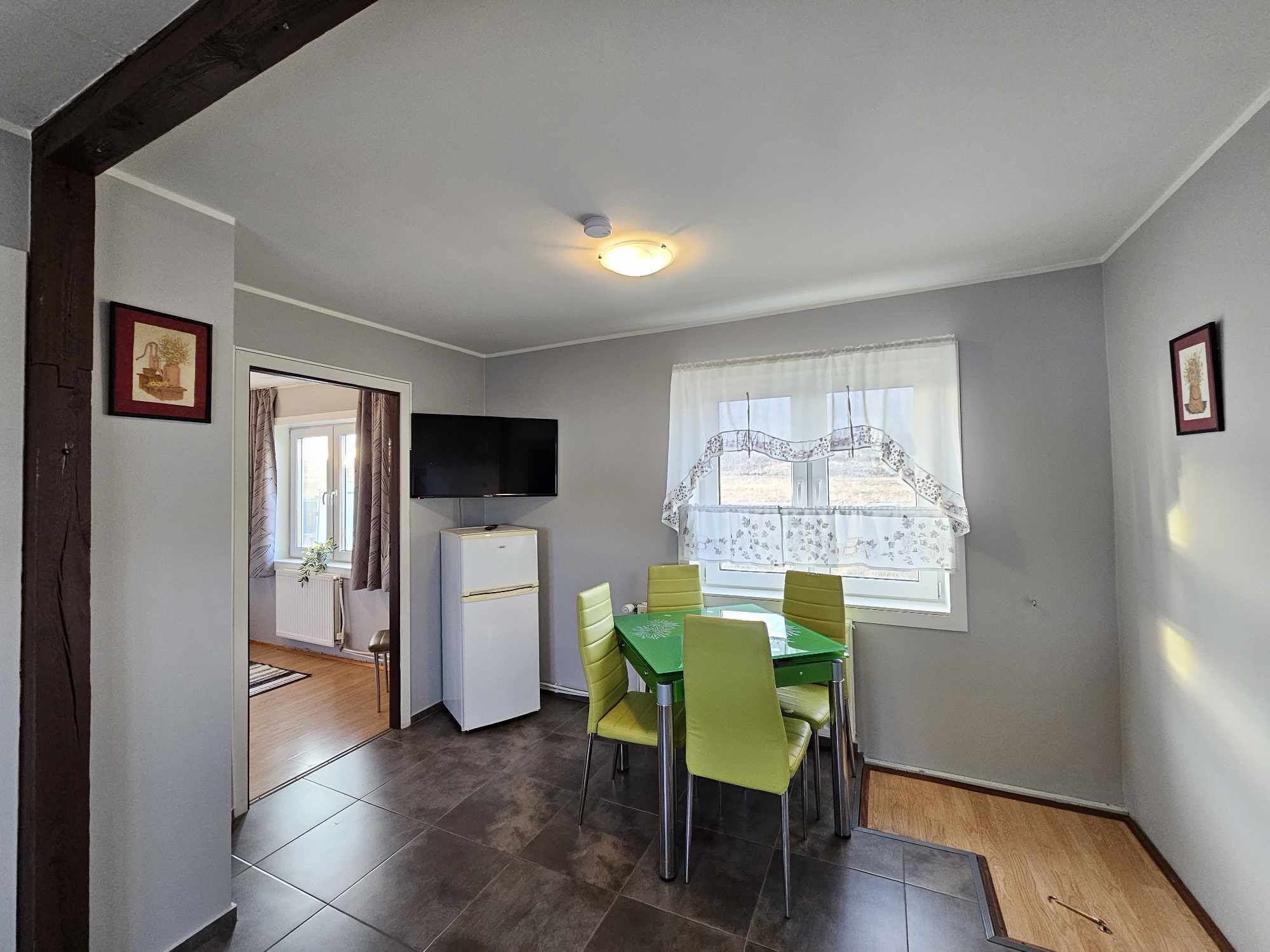 A compact kitchen with a dining area, a TV mounted above the counter, and a window with sheer curtains.