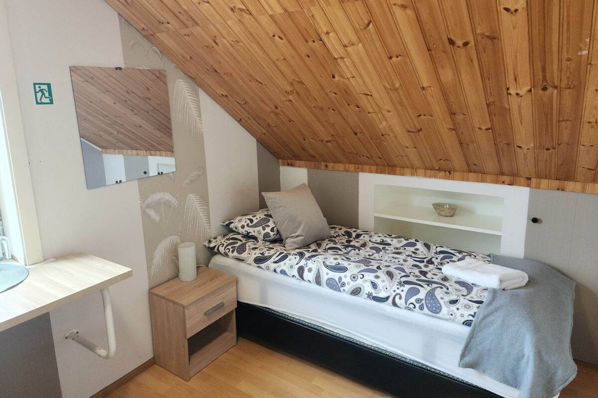 A small, cozy bedroom with a sloped ceiling, a bed with patterned bedding, a bedside table, a mirror, and a shelf with a bowl.