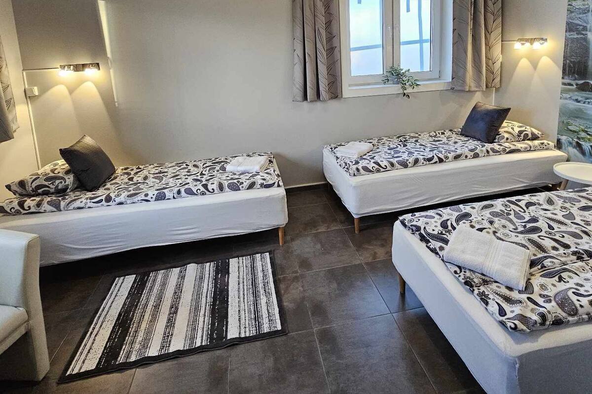 A room with two single beds, a small table, a chair, a window with curtains, and a wall-mounted light fixture.