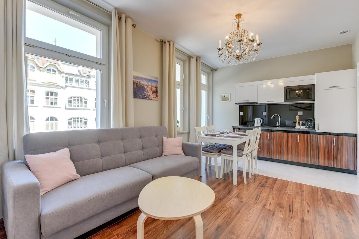 A modern living room with a grey sofa, a round table, and a kitchen area with white cabinets and a black microwave.