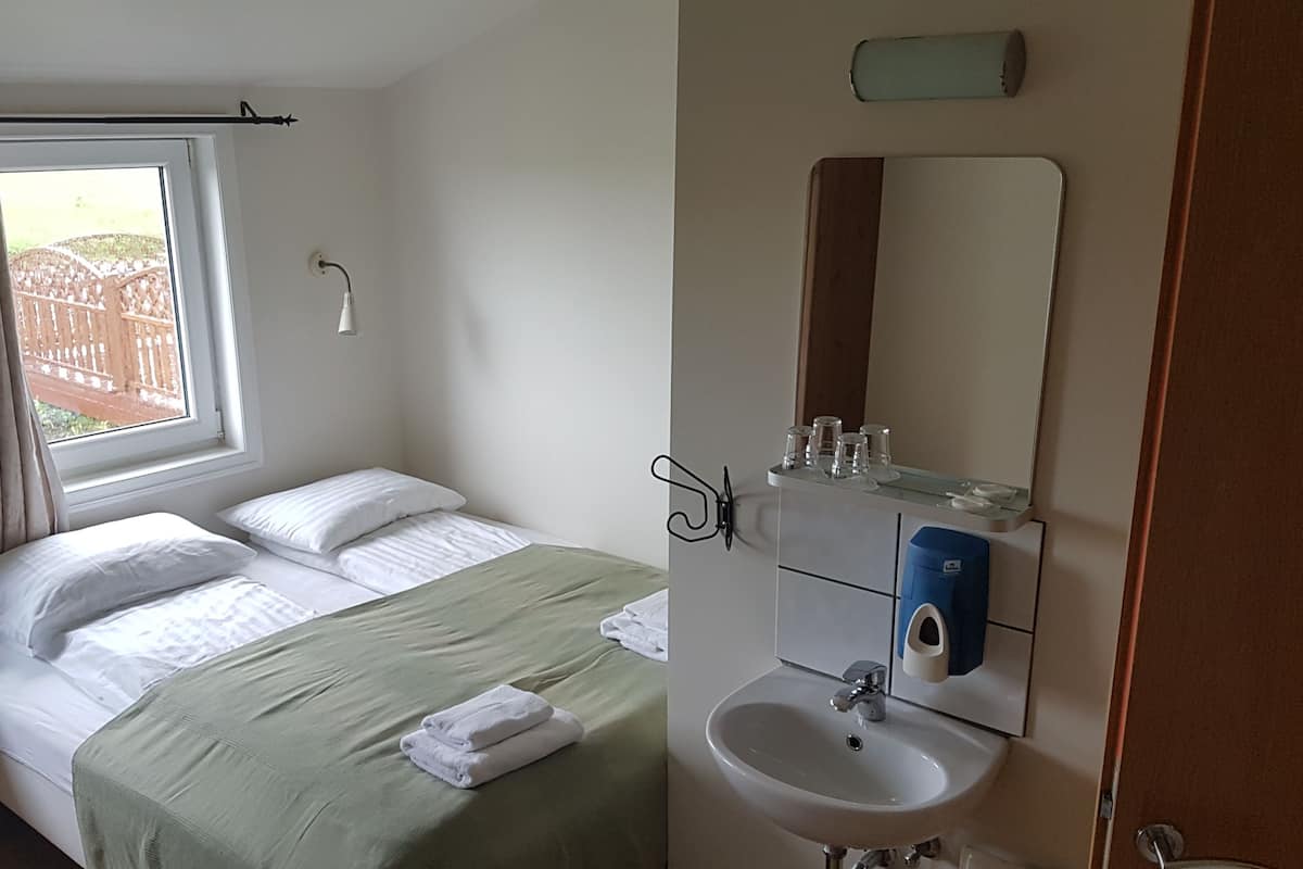 A bedroom with a bed, a window with curtains, a bedside table, a wall-mounted mirror, and a sink.