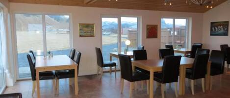 A dining area with wooden tables and chairs, large windows with a view of the outdoors, and a wooden ceiling with exposed beams.