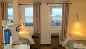 Habitación de hotel con baño, dos camas, ventana con vista al mar y una mesita con lámpara.