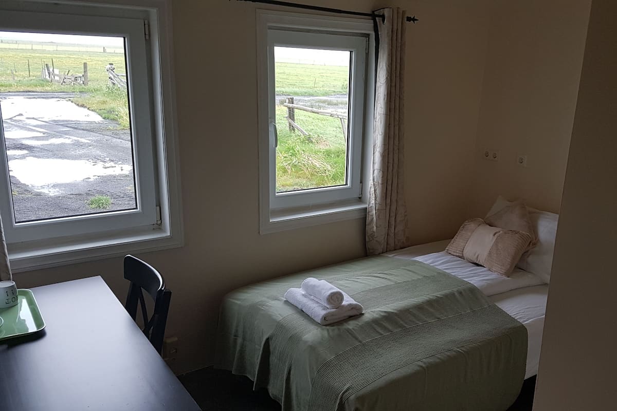 A small, simple room with a bed, a table, and two windows.