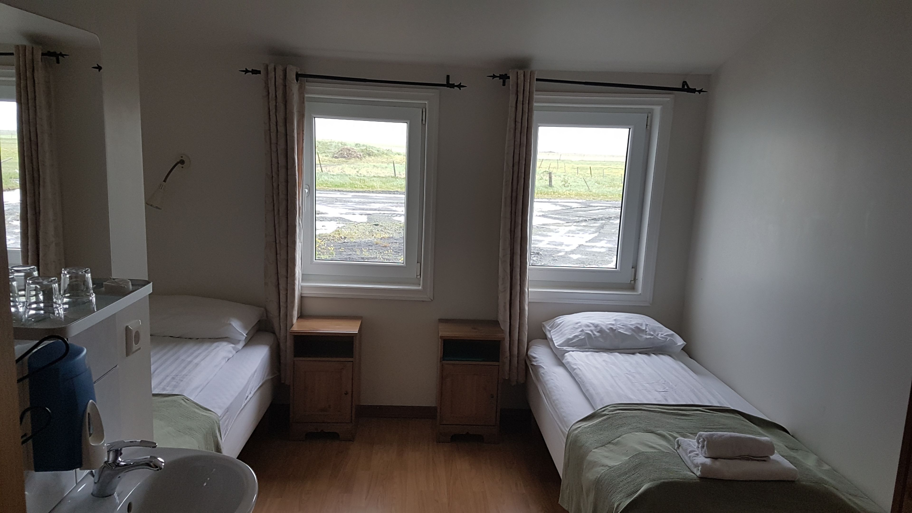 Una habitación pequeña y bien iluminada con dos camas individuales, un lavamanos y un espejo.