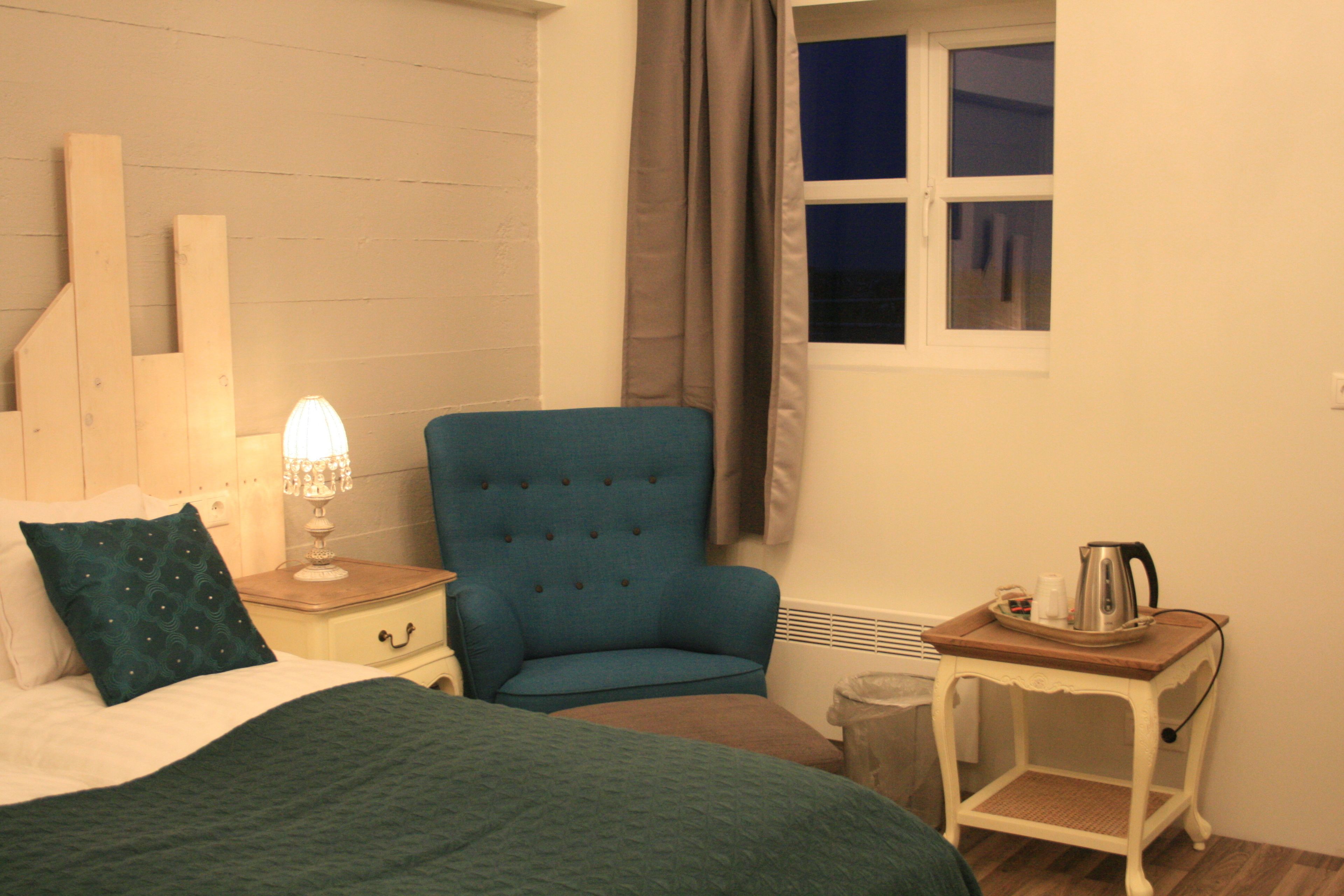 Un dormitorio con cama, un sillón azul, una mesita de noche con lámpara, una ventana con cortinas y una pequeña mesa con hervidor.