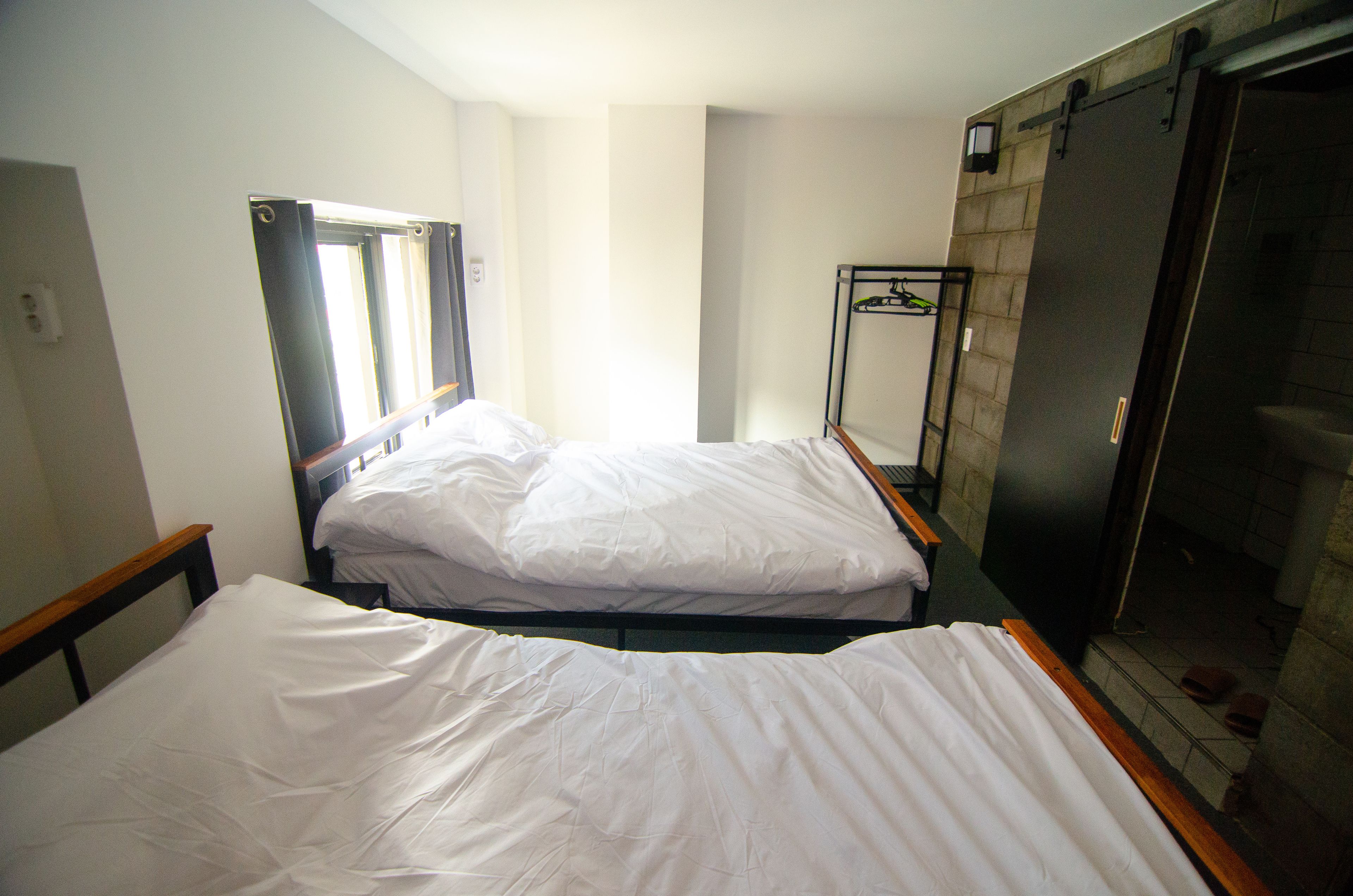 Dos camas individuales con cabeceros de madera y sábanas blancas en una habitación con piso de baldosas y un ventanal pequeño.