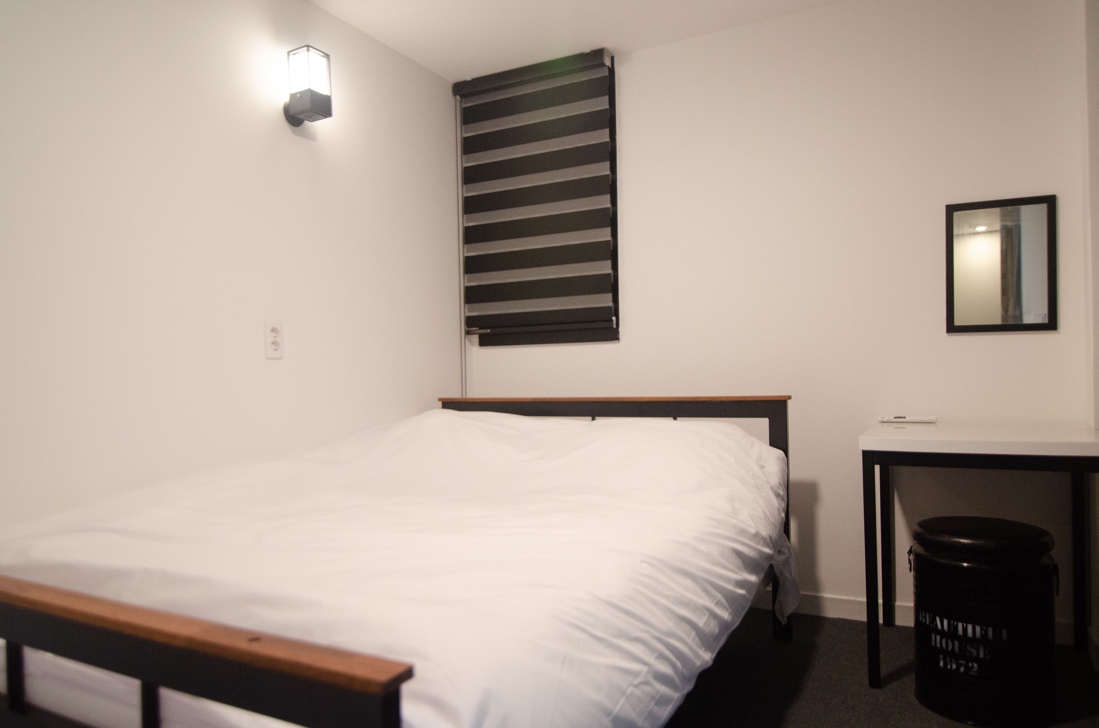 A bedroom with a bed, a desk, a chair, a trash bin, a wall-mounted light, and a window with blinds.