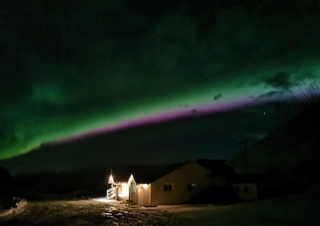 A house with a lit porch under a green aurora display.