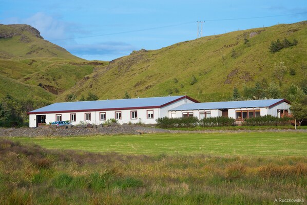 A white building with a red roof, situated in a grassy field with a hilly backdrop. - Hótel Búrfell (Vik I Myrdal)
