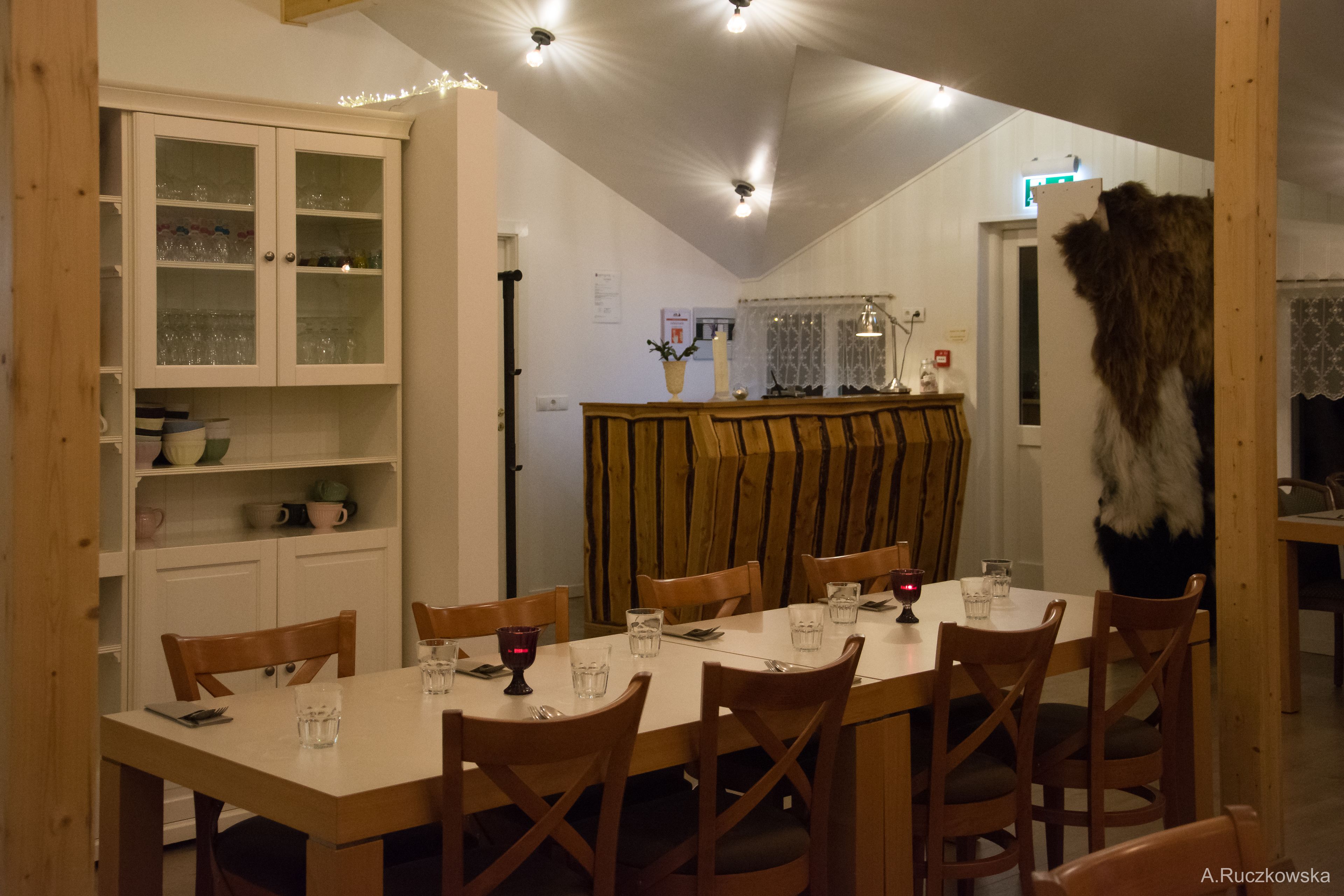 Un coin repas avec une longue table dressée pour six personnes, entourée de chaises en bois. Un buffet avec des portes en verre se trouve à gauche, et une fenêtre recouverte d’un rideau se trouve derrière la table.