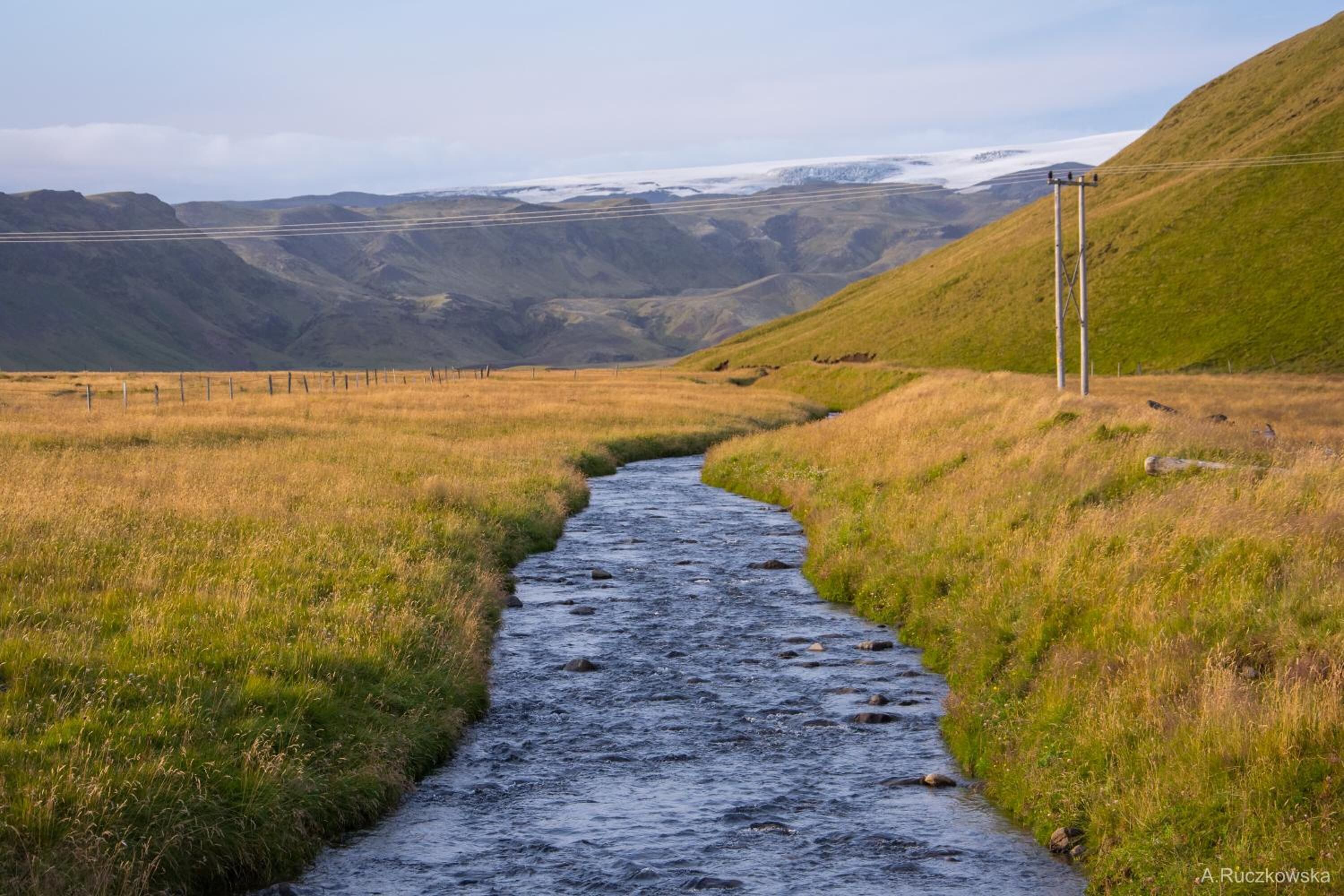 A narrow stream flowing through a grassy valley with hills in the background and a power line structure on the right.