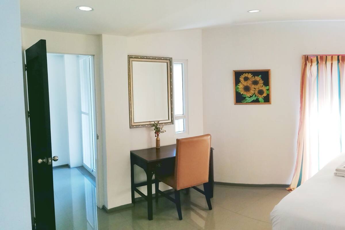 A hotel room with a bed, a desk with a chair, a lamp, a window with curtains, and a painting on the wall.