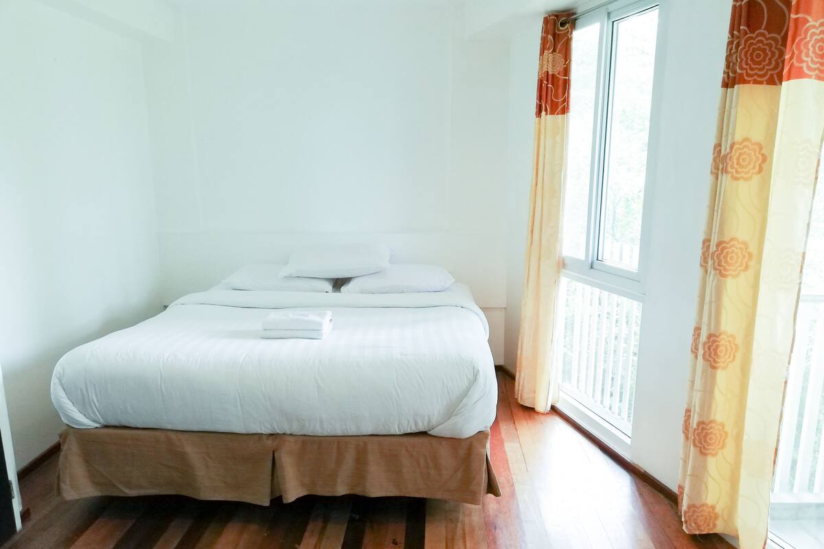 A neatly made bed with white linens and a folded towel, wooden flooring, and a window with curtains.