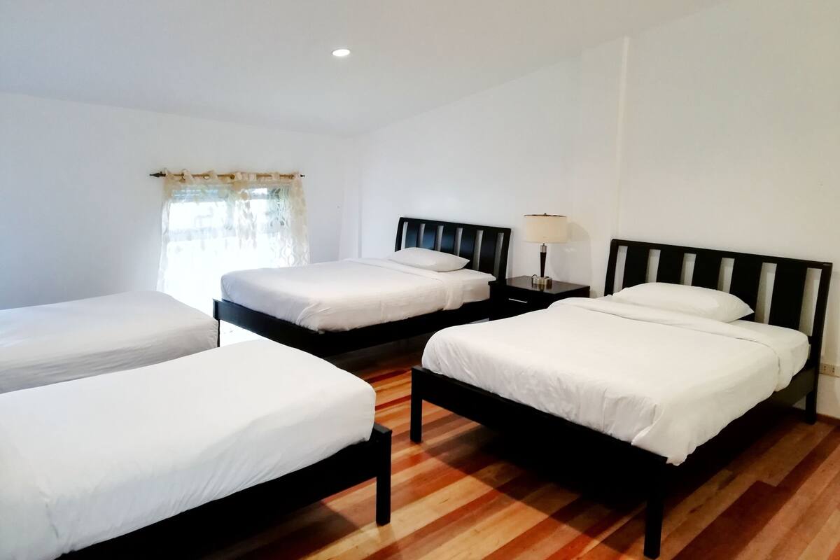 A hotel room with three single beds, wooden flooring, white walls, and a window with a curtain.