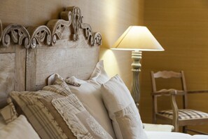 A neatly made bed with multiple pillows and a headboard with carved details. A lamp and chair are visible in the background.