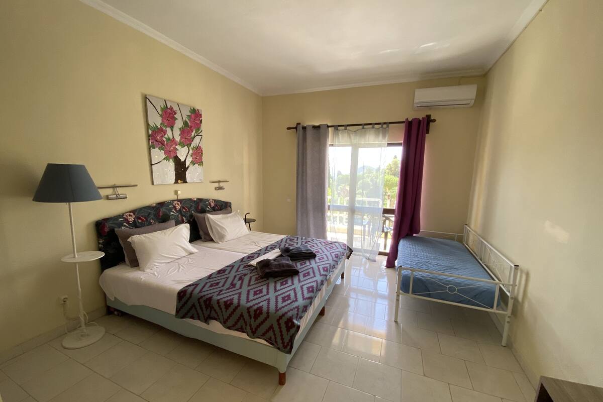 A hotel room with a large bed, a smaller bed, a floor lamp, a painting on the wall, and a view of the outdoors through the window.