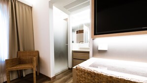 A room with a wicker chair, a large mirror, a flat-screen TV, and a wooden floor. - Hotel Corona (Caprino Veronese)