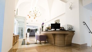 A modern reception area with a curved wooden desk, a purple ottoman, and a chandelier. - Hotel Corona (Caprino Veronese)