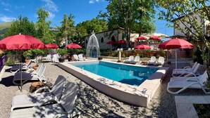 A pool area with lounge chairs and umbrellas, surrounded by trees and a stone building. - Hotel Corona (Caprino Veronese)