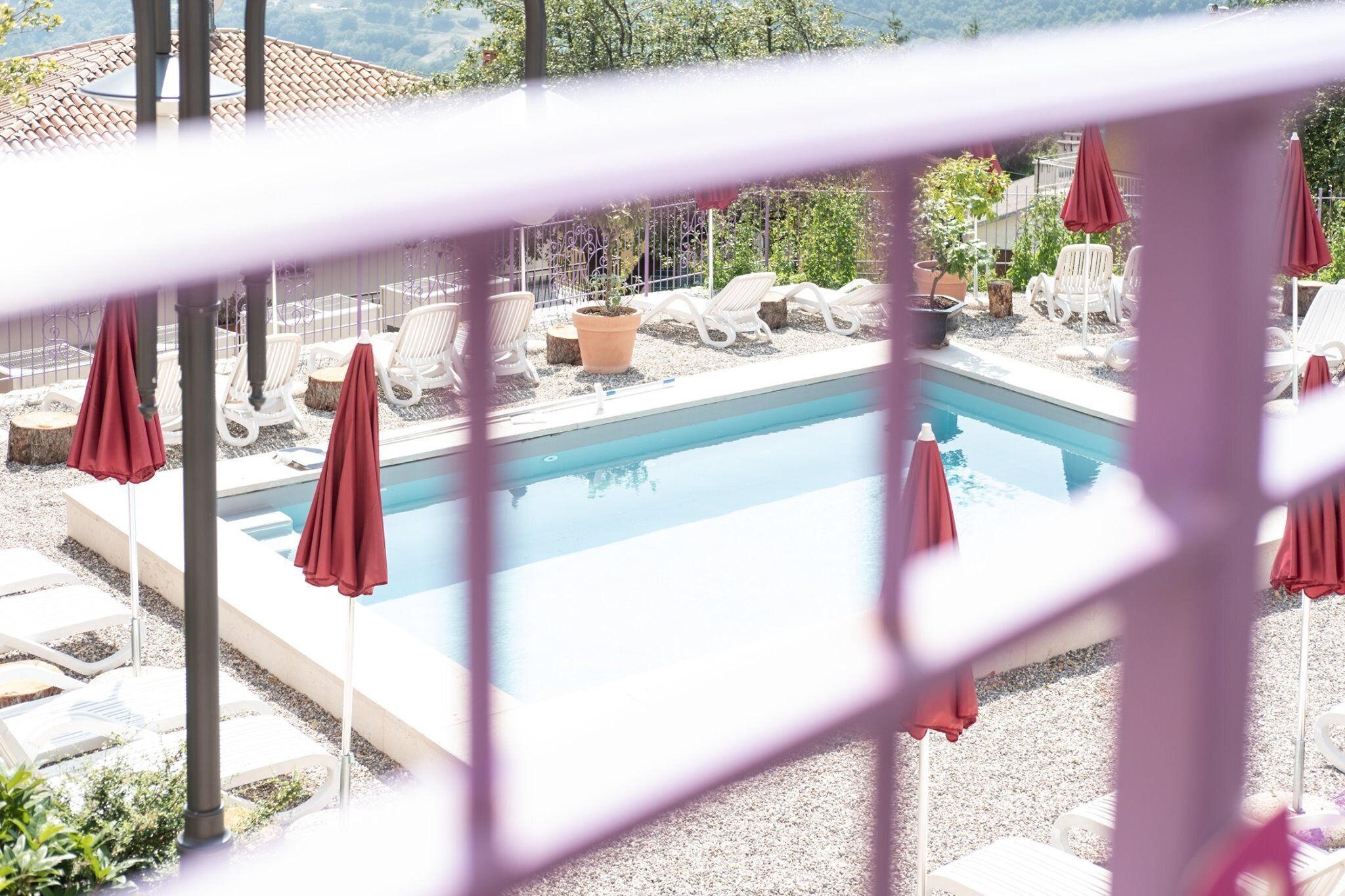 Zona junto a la piscina con sillones de descanso y sombrillas, rodeada por una valla y follaje.