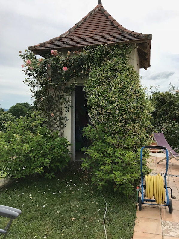 A small, rustic gazebo with a tiled roof, surrounded by lush greenery and flowering plants.
