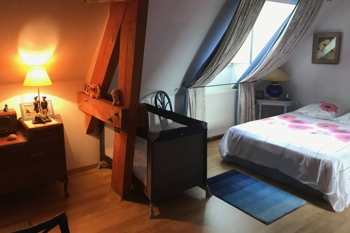A bedroom with a sloped ceiling, a wooden bed frame, a bedside table with a lamp, a window with curtains, and a small chair.