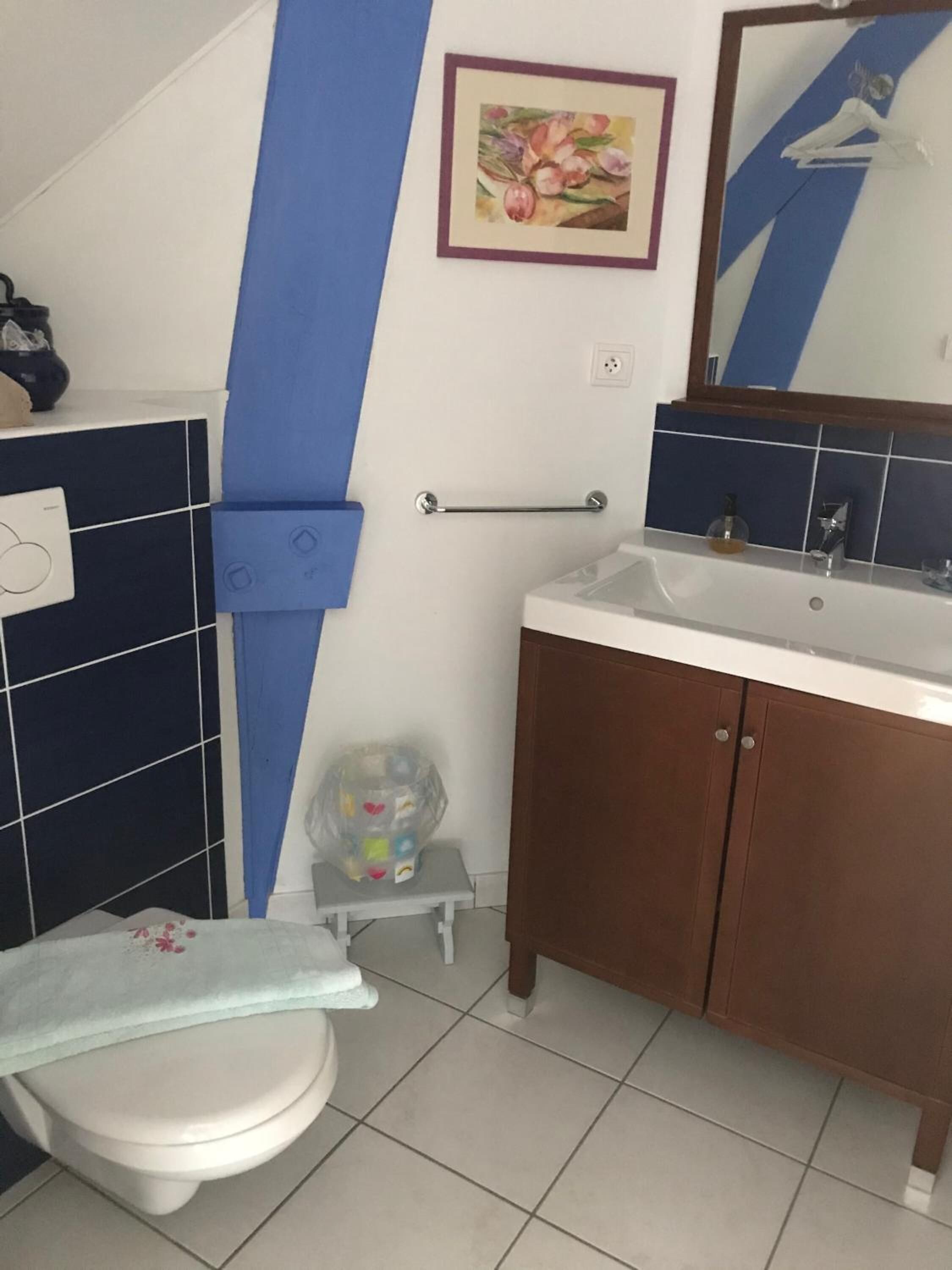 A bathroom with a toilet, sink, and cabinet. There is a blue towel hanging on the wall and a small stool with a plastic bag on it.