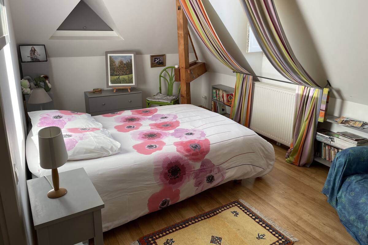 A bedroom with a bed, a nightstand, a lamp, a chair, a radiator, and a window with curtains.