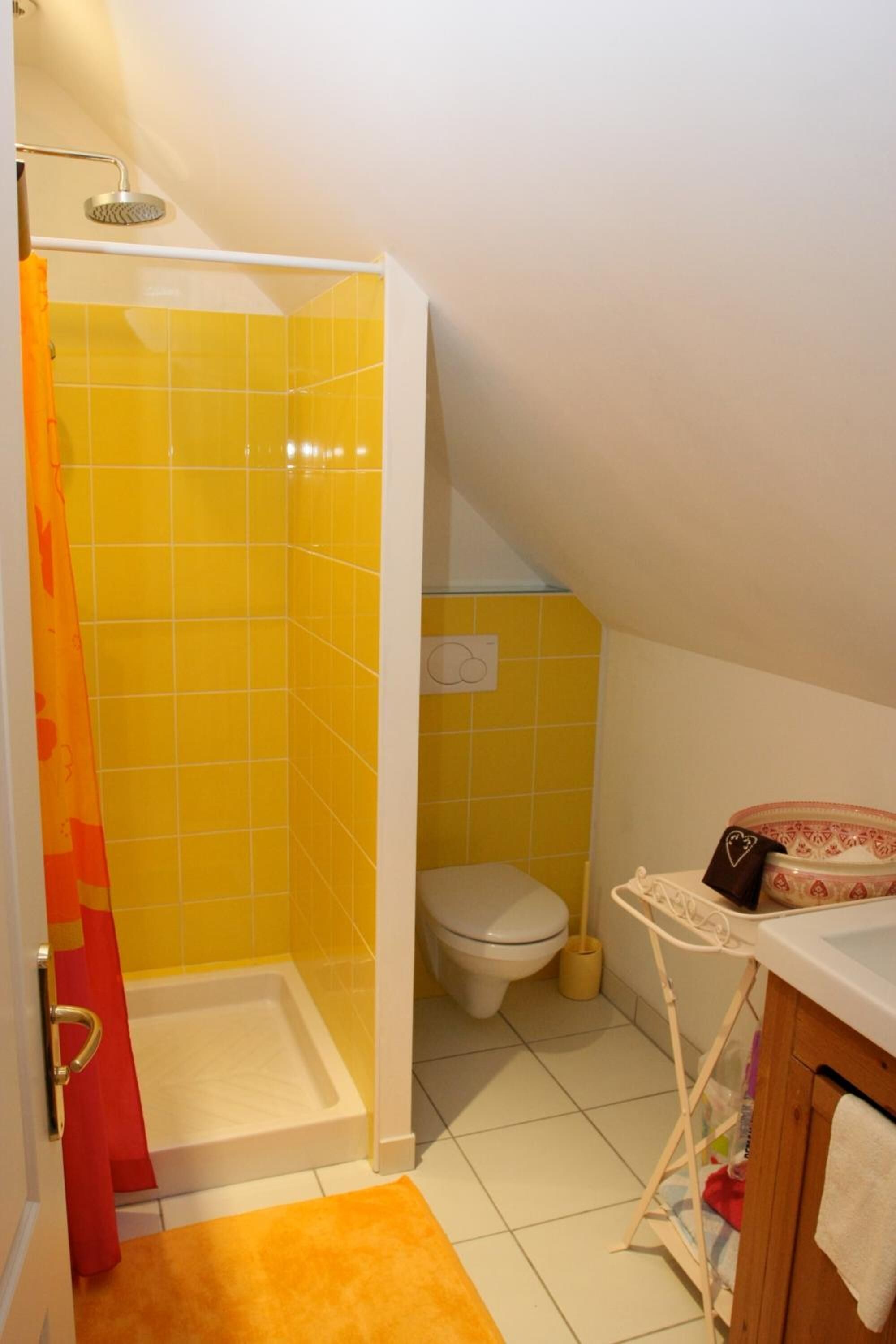 Casa de banho com azulejos amarelos, chuveiro, sanita e uma pequena mesa com uma toalha.