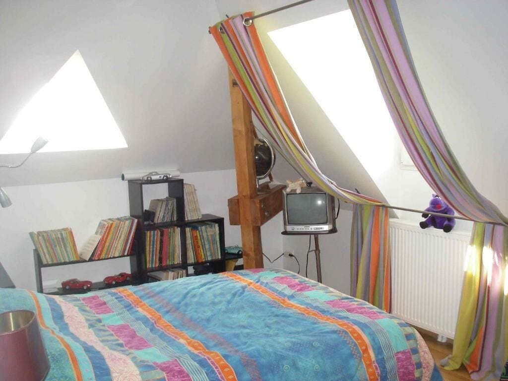 Un dormitori amb un llit, una prestatgeria, una televisió i una finestra amb cortines.