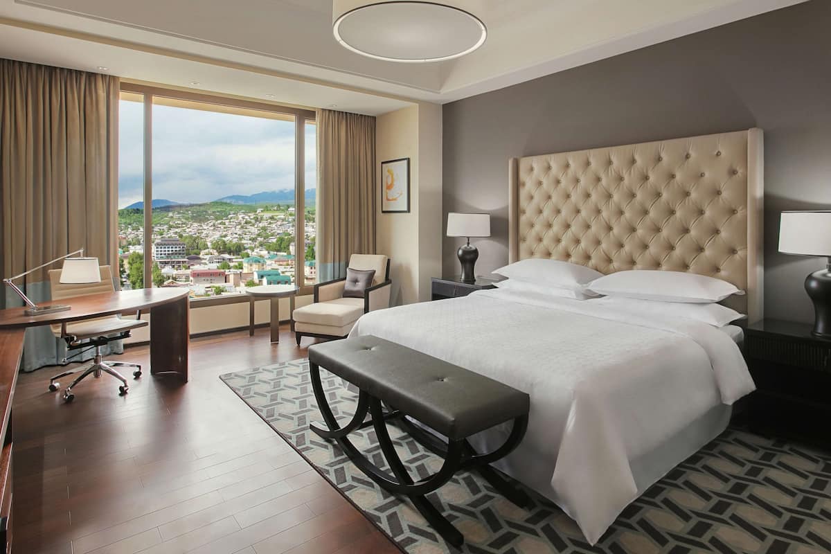 A hotel room with a large bed, a desk, a chair, and a view of a cityscape.
