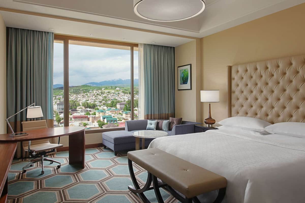 A hotel room with a large bed, a sofa, a desk, and a view of a cityscape.
