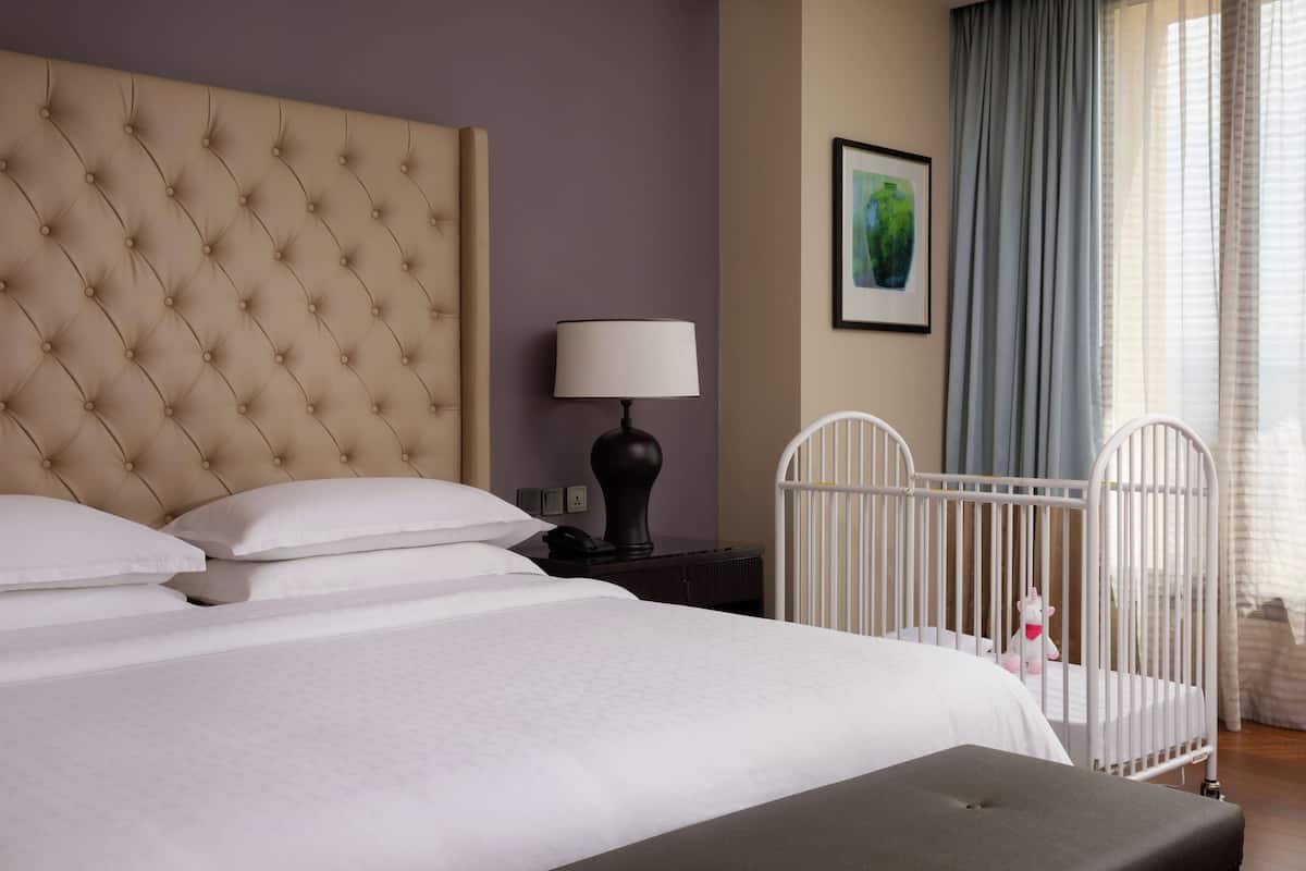 A neatly made bed with white linens, a tufted headboard, a bedside lamp, and a crib with a white railing.