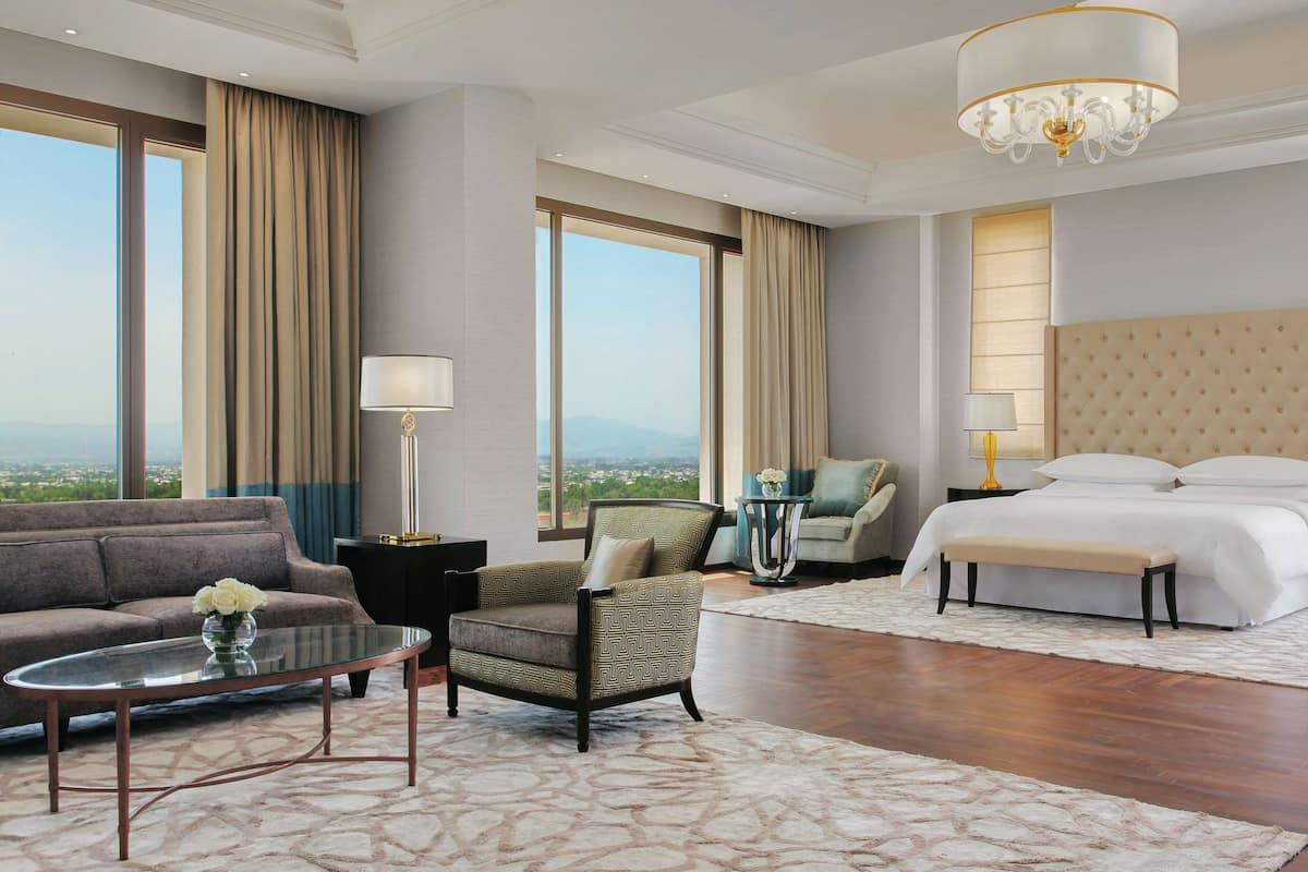A hotel room with a large bed, a sofa, a chair, a coffee table, and a view of the city.