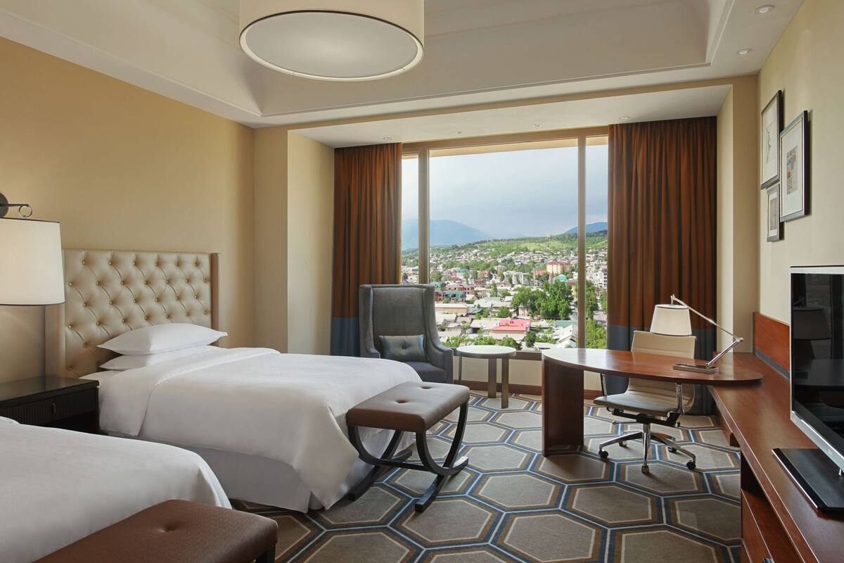 A hotel room with two beds, a desk, a chair, a TV, and a large window offering a city view.