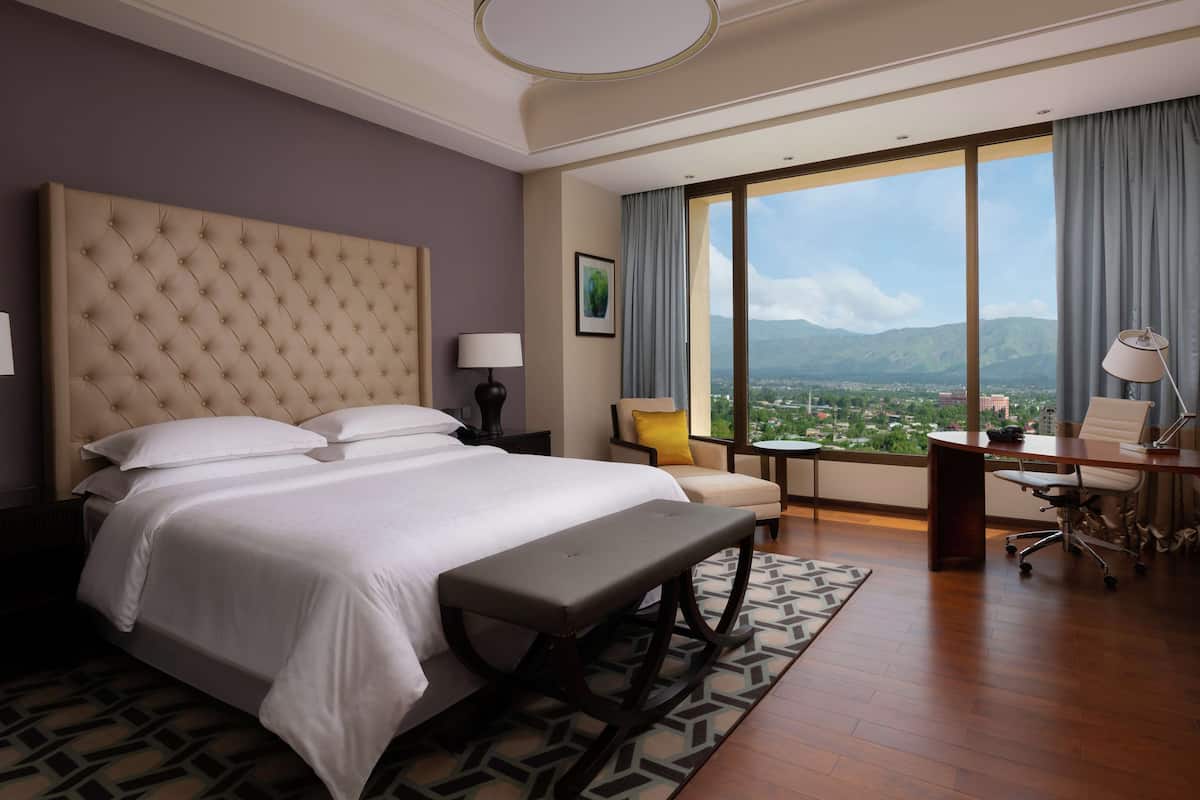 A hotel room with a large bed, a desk, and a view of a cityscape through large windows.