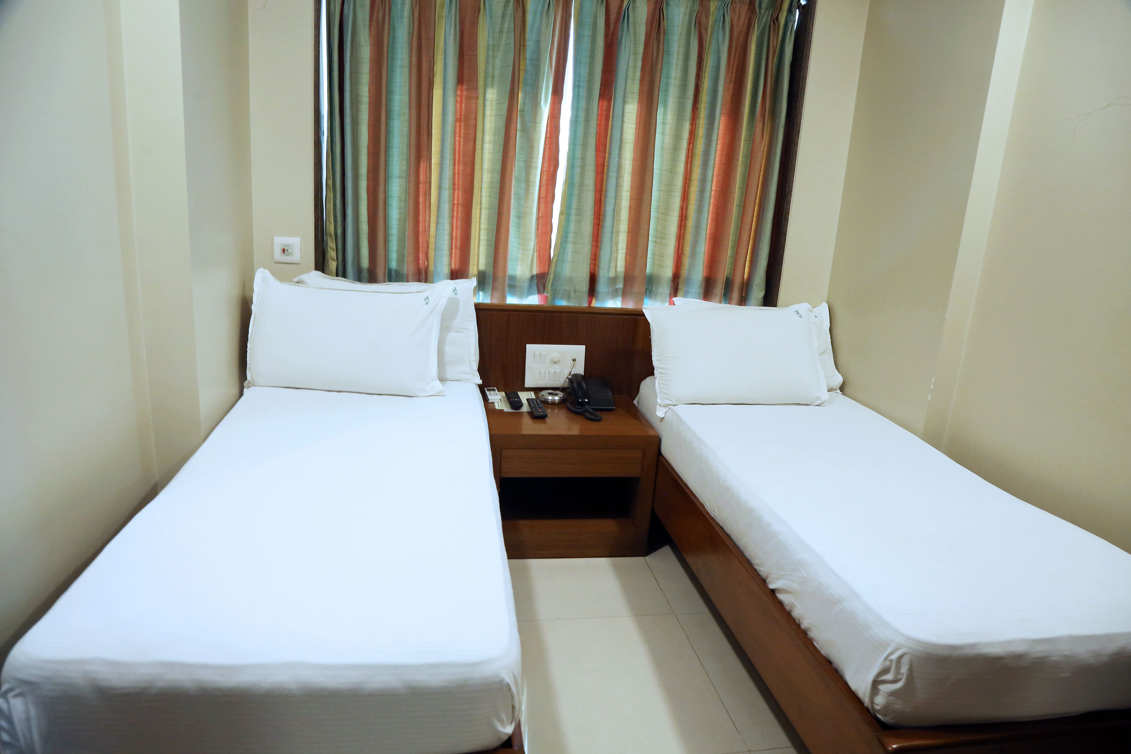 A small, clean room with two single beds, a wooden nightstand with a phone and remote controls, and a window with colorful curtains.