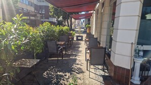 Un área de café al aire libre con mesas y sillas, rodeada de plantas y la fachada de un edificio.