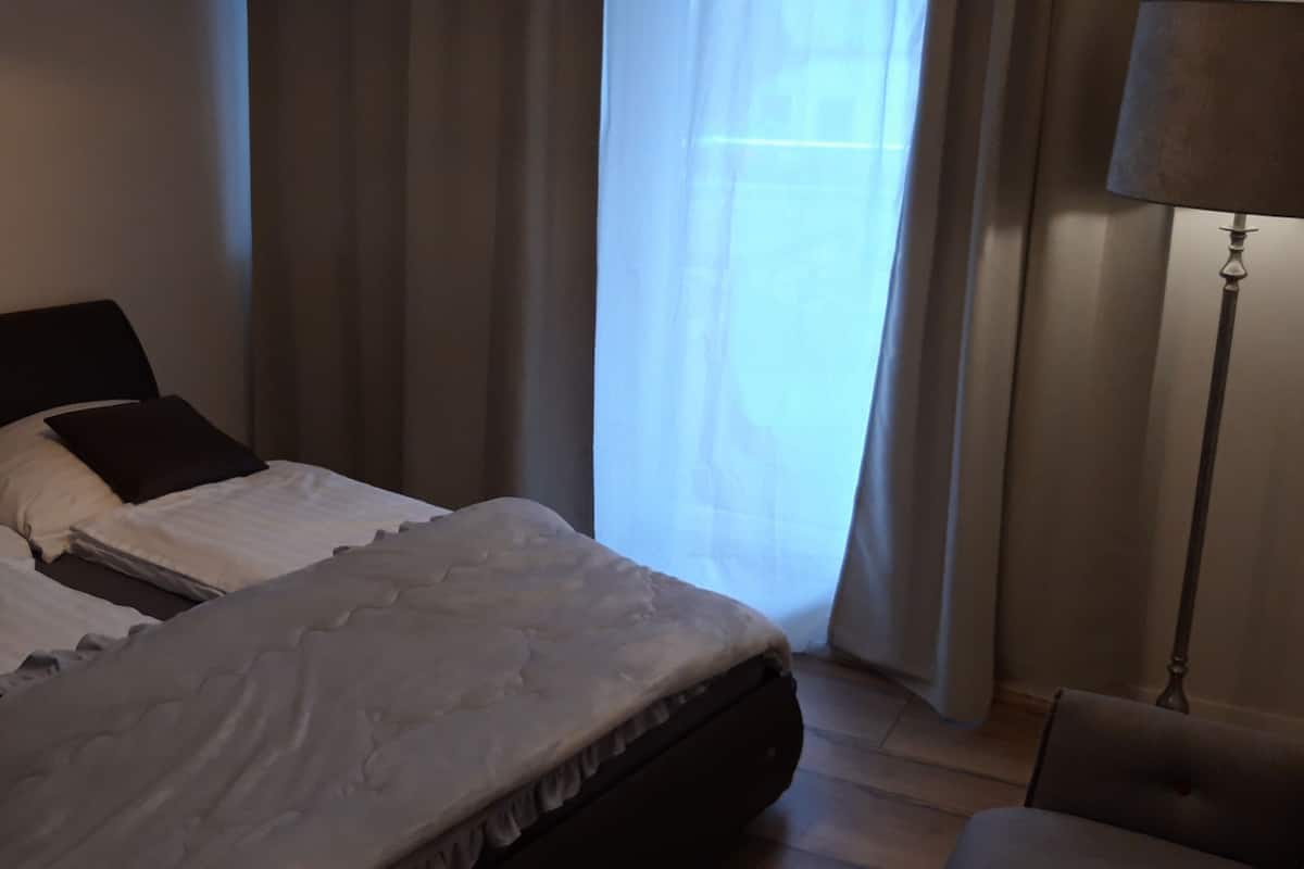 A neatly made bed with white linens and dark pillows, a bedside lamp, and a chair in a room with a large window and curtains.