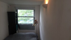 A small, narrow room with a single bed, a window with a view of trees, and a wall-mounted light fixture. - The Portobello Loft (London)