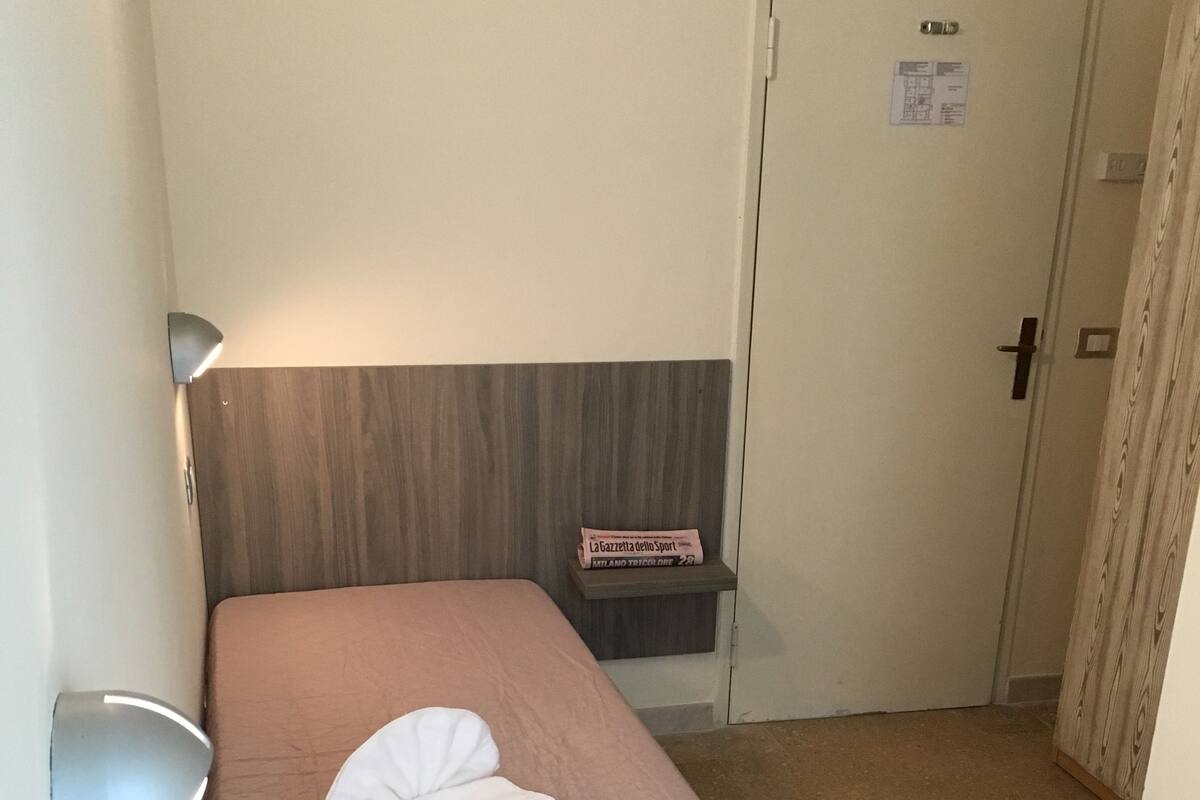 A single bed with a headboard, a bedside table with a lamp, a small shelf with magazines, and a door with a sign.