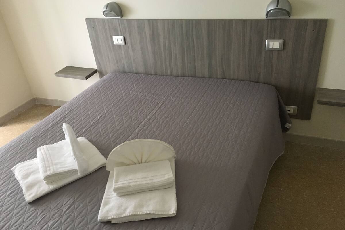 A neatly made bed with a grey quilt and two neatly folded white towels.