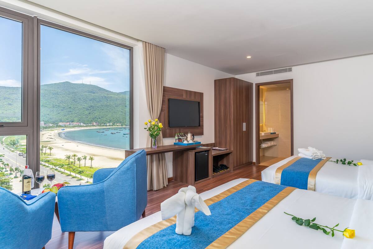 Ein Hotelzimmer mit großem Fenster mit Blick auf einen Strand, einem an der Wand befestigten Fernseher und zwei Betten mit Handtüchern und einer Blume.
