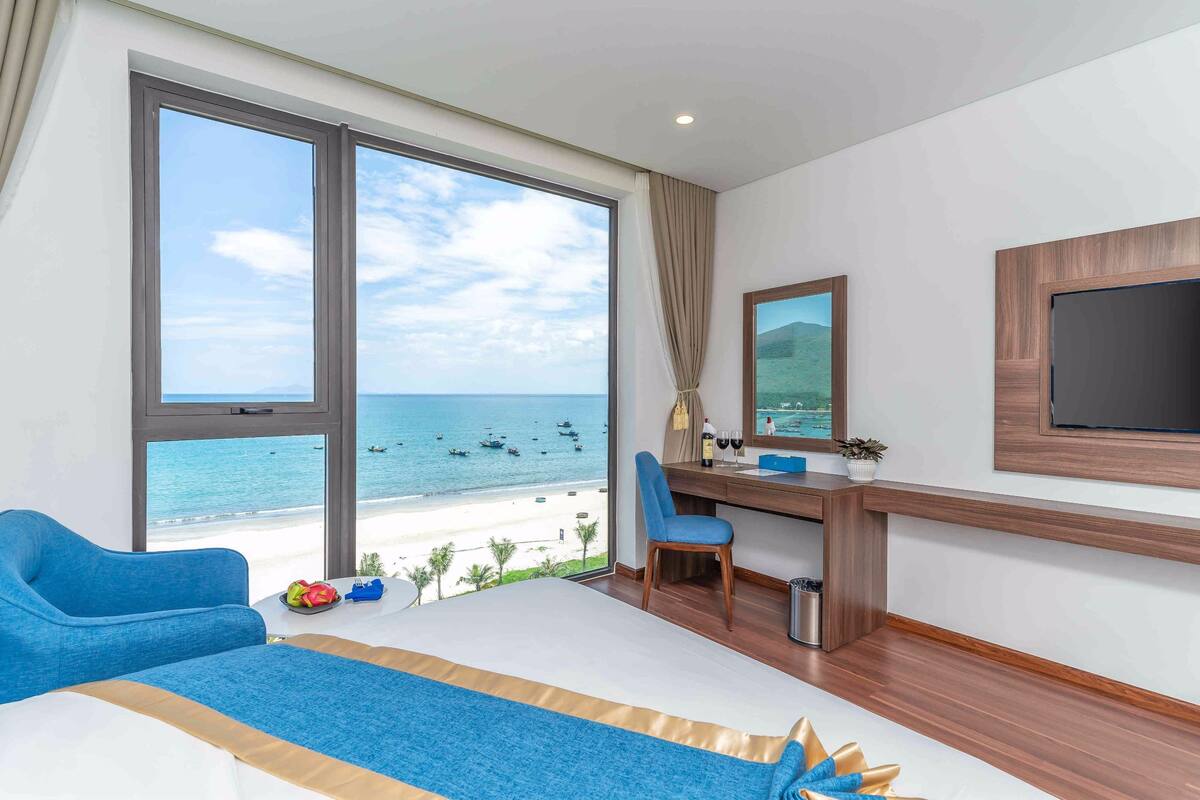 Ein Hotelzimmer mit großem Fenster und Blick auf den Strand, einem Bett, einem Schreibtisch mit Stuhl und einem Wand-Fernseher.