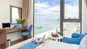 Une chambre d’hôtel dotée d’une grande fenêtre donnant sur une plage, d’un bureau avec un ordinateur portable, d’une chaise et d’un lit avec une serviette et des fruits à proximité.