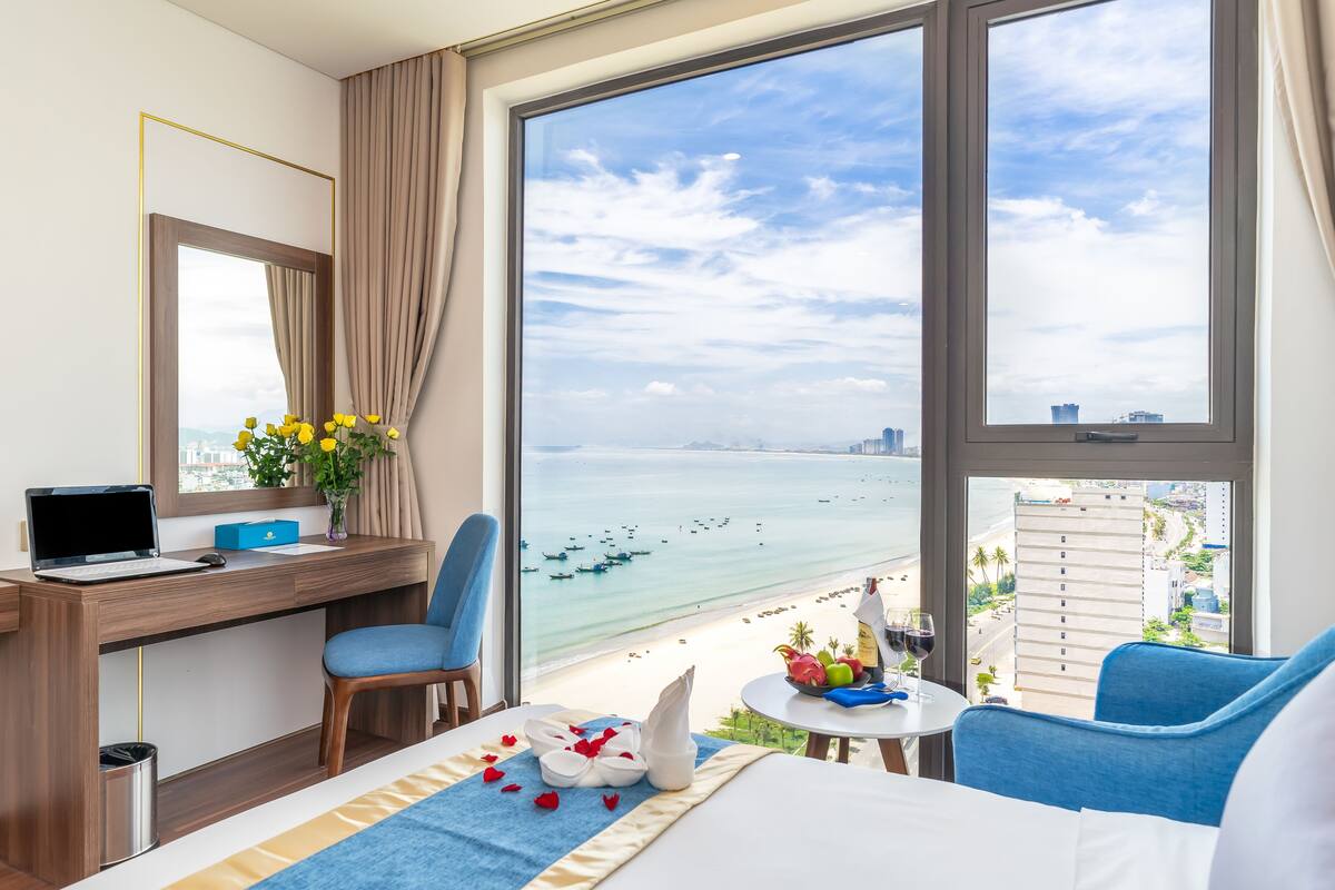 Ein Hotelzimmer mit großem Fenster, das einen Blick auf einen Strand bietet, einem Schreibtisch mit einem Laptop, einem Stuhl und einem Bett mit Handtuch und Früchten auf dem Nachttisch.