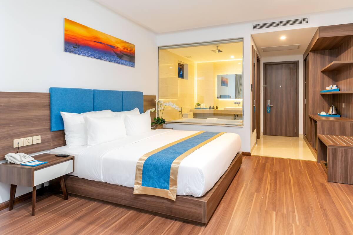 Ein Hotelzimmer mit einem großen Bett, einem Schreibtisch, einem Fernseher und einem durch eine Glastür einsehbaren Badezimmer.